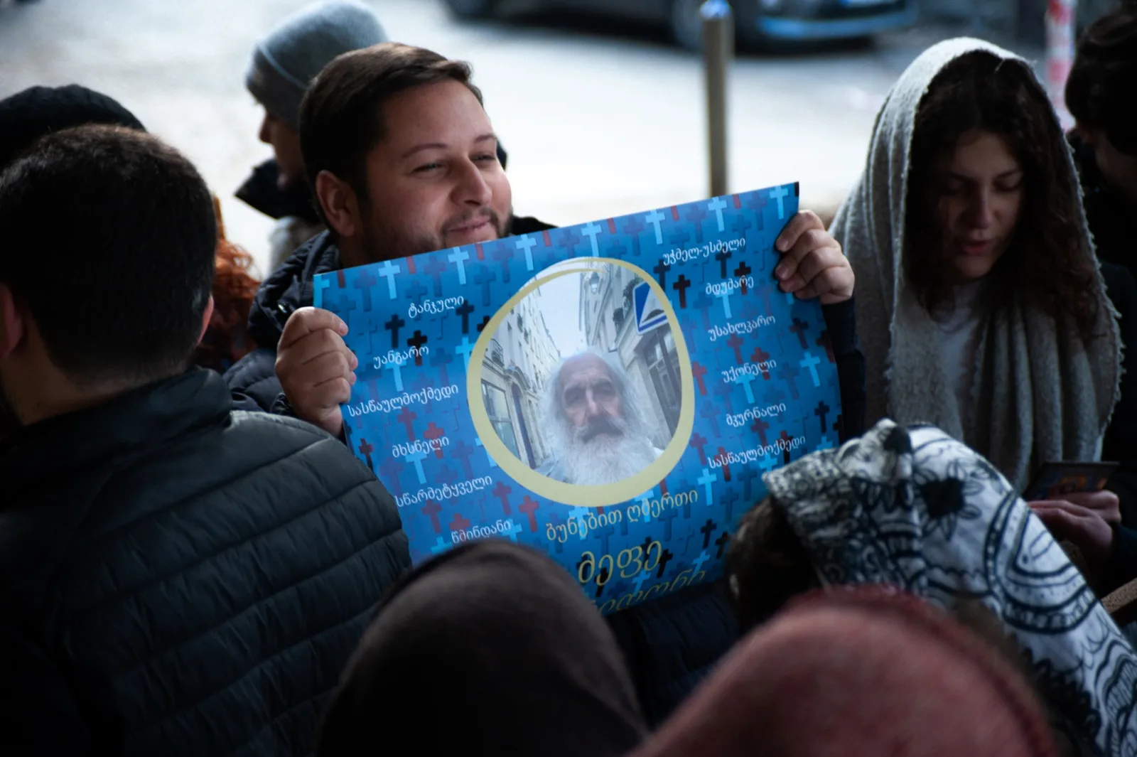 Holy Smear still: a man in a crowd holding up a poster of a bearded elder ringed with crosses