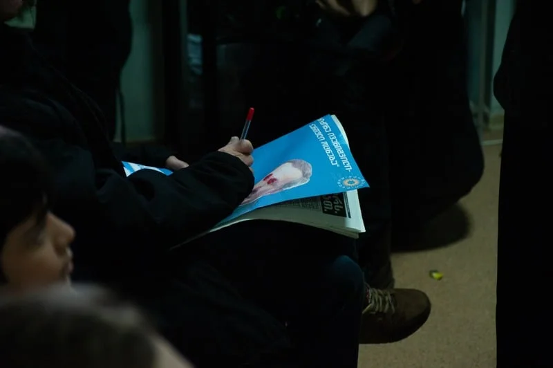 Holy Smear still: a hand signing a blue poster in a crowd