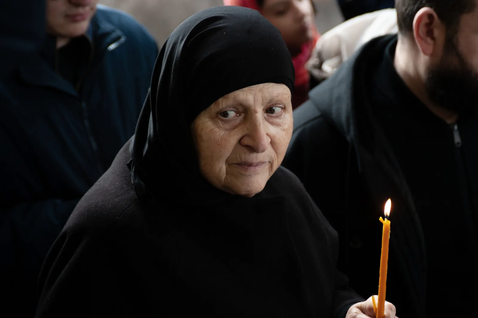 Holy Smear still: an older woman in a black headscarf holding a candle in a crowd