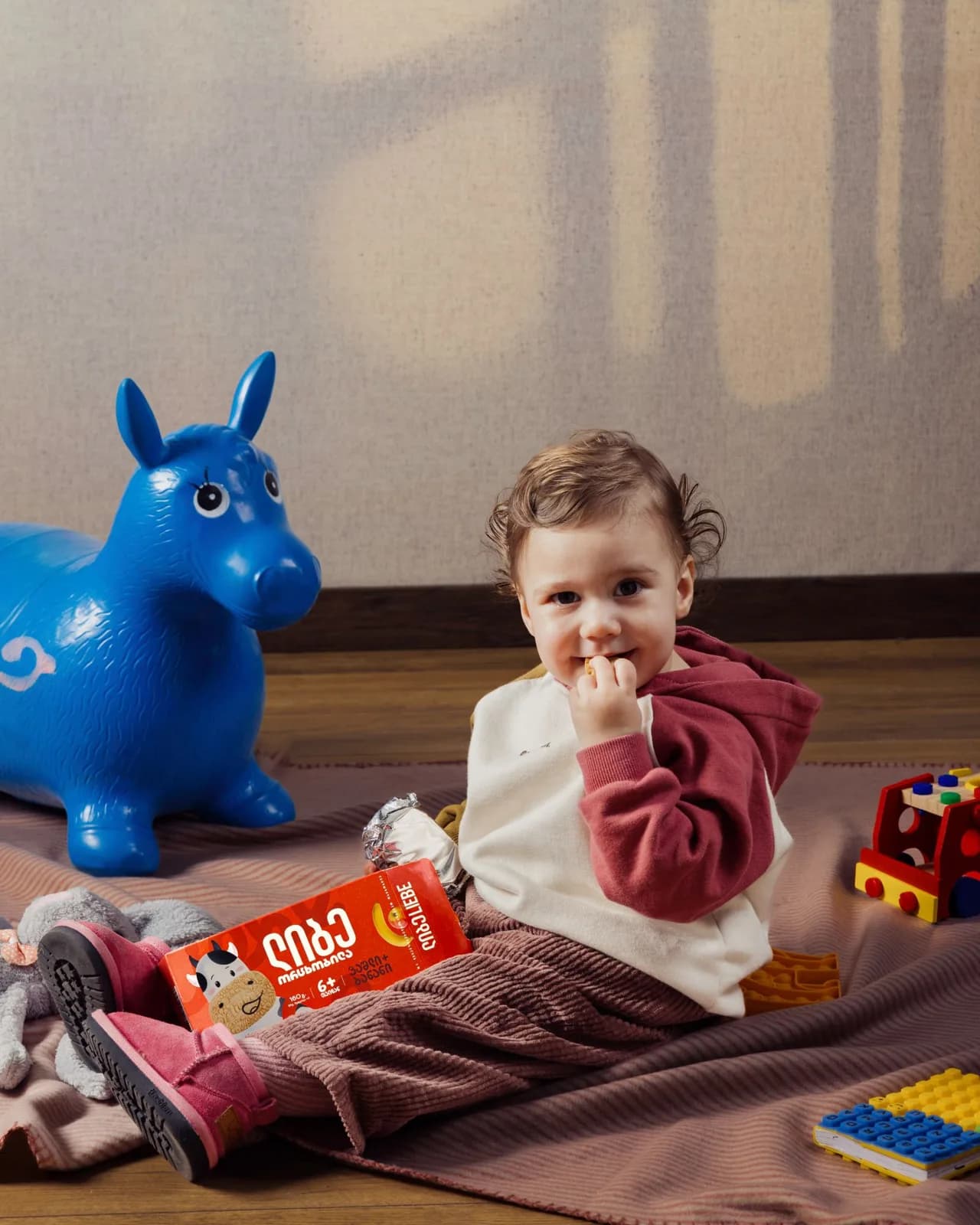 Toddler on a blanket eating a biscuit next to a blue toy horse and a Liebe pack