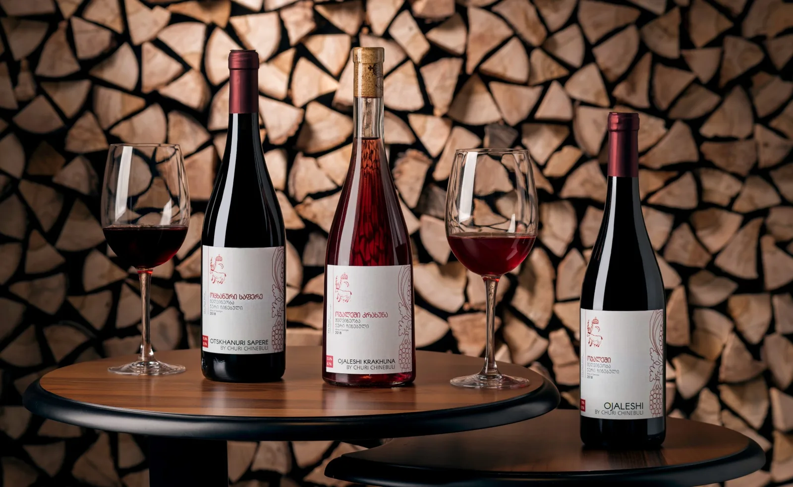 Wine ad: three Churi Chinebuli bottles and glasses on a table in front of a stacked-wood wall